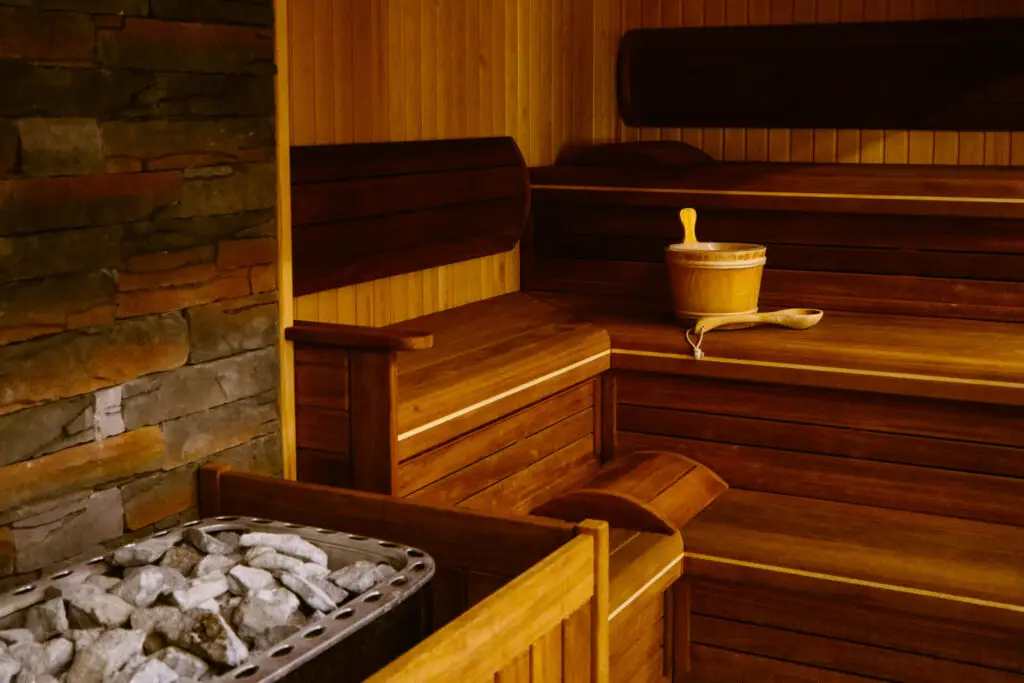 Innenraum einer traditionellen Sauna mit Steinofen, Holzbänken und einem Holzeimer mit Kelle.