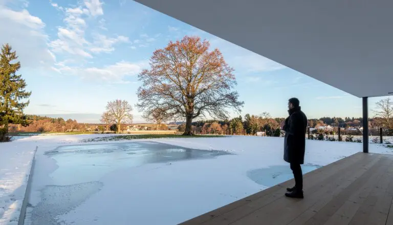 Gefrorener Pool mit Schneeschicht vor moderner Terrasse und freistehendem Baum im Winterlicht.